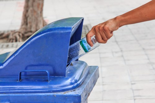A personâ€™s hand is seen placing a clear plastic bottle into a blue recycling bin with a hinged lid, situated outdoors on a paved walkway. The person's arm is extended from the right side of the image, with their hand grasping the bottle by its middle section. The recycling bin is made of plastic with a matte finish, featuring a slightly rounded lid that is partially open, revealing the interior. In the background, a tree trunk and a paved surface are visible, indicating a public or urban environment. The scene emphasizes waste separation and recycling practices, relevant to rubbish removal services in Croydon, with the focus on proper disposal of plastic bottles in designated recycling containers.